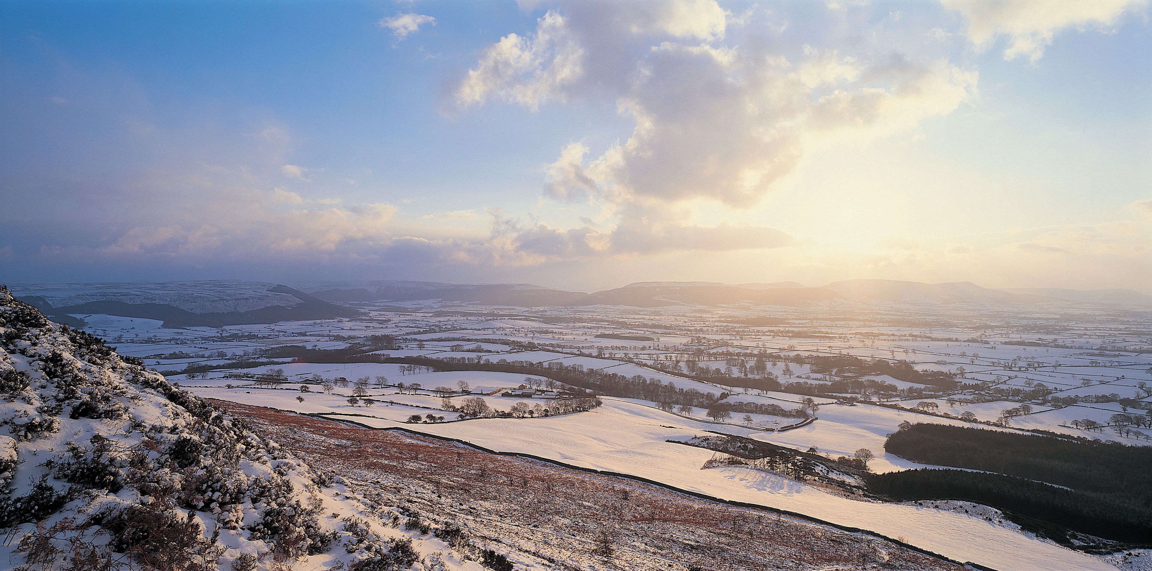Paisagem coberta de neve, com colinas onduladas e uma vista distante de um vale.