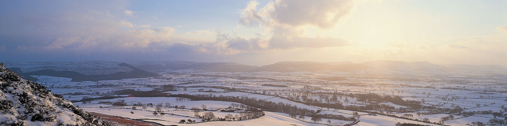 Paisagem coberta de neve, com colinas onduladas e uma vista distante de um vale.