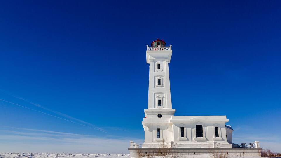 A white lighthouse with a red top, surrounded by snow-covered landscape under a clear blue sky.