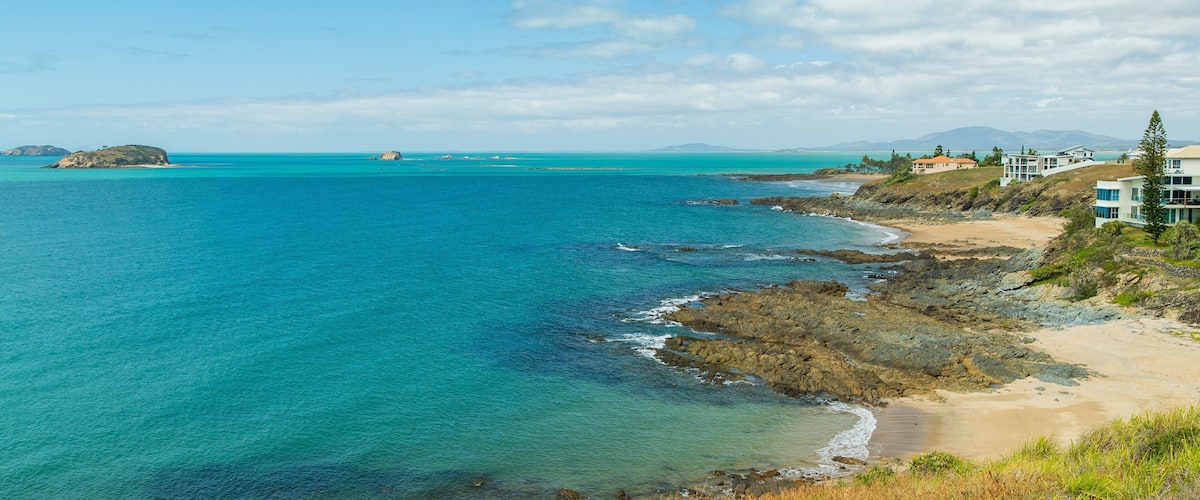 A coastal landscape with clear turquoise waters, rocky shores, and modern buildings on the right.