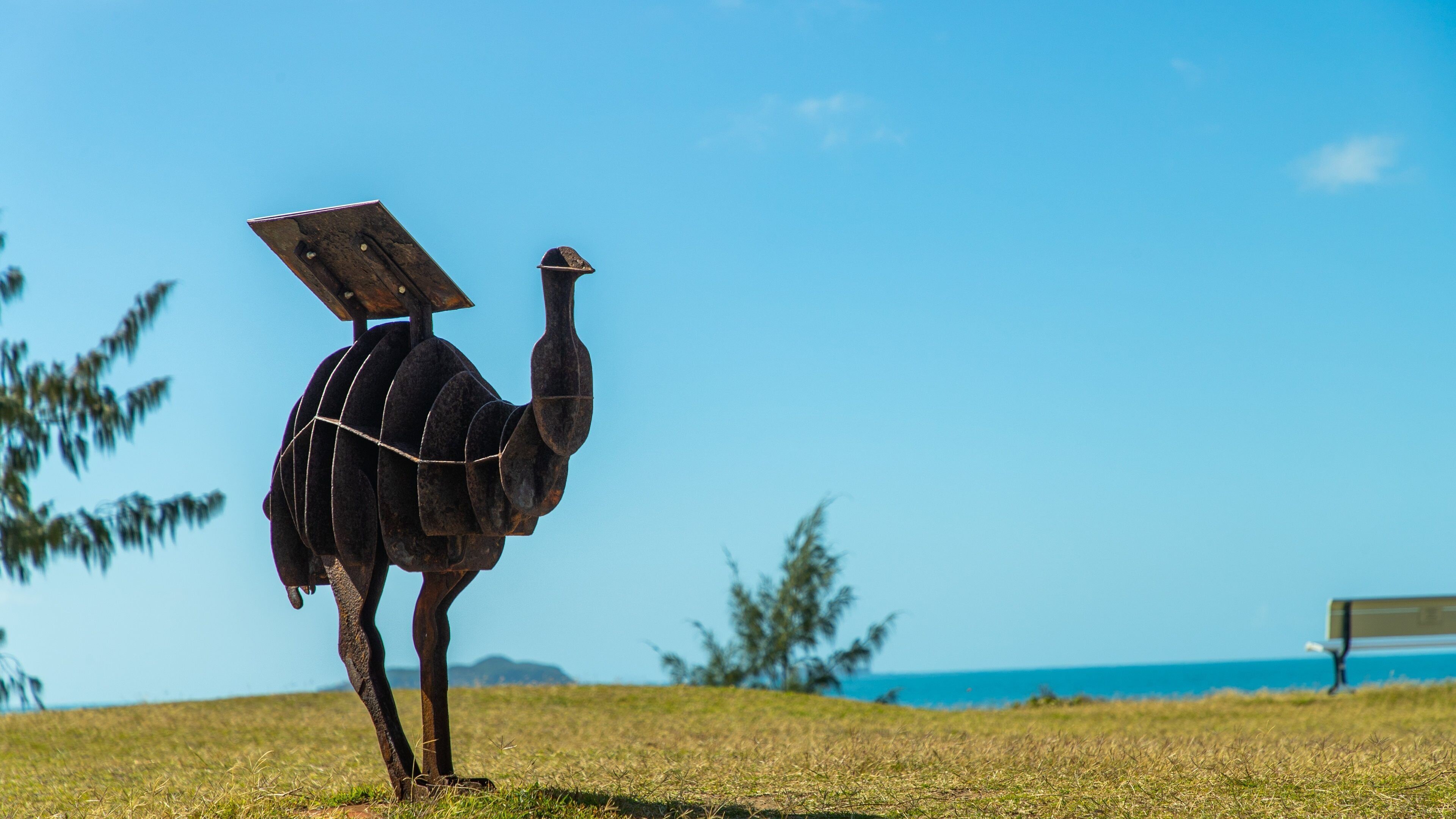 Una escultura de avestruz con un panel solar en el lomo, ubicada en un campo verde frente a un cielo azul despejado y el océano.