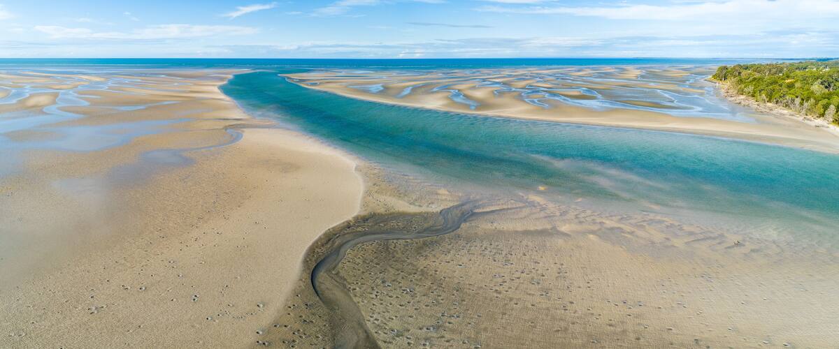 A coastal landscape with a wide sandy beach, a clear blue sea, and a narrow water channel.