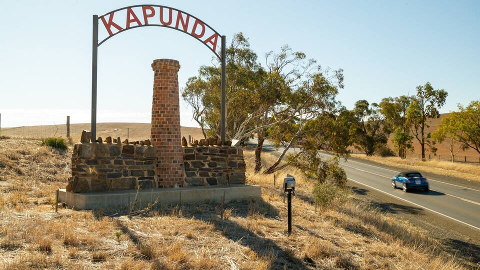 A sign reading "Kapunda" with a stone pillar and a brick chimney, set against a backdrop of a road, dry grass, and trees.