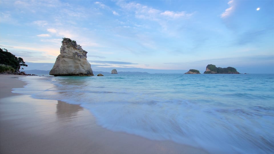 A beach with calm waters, a large rock formation, and distant islands.