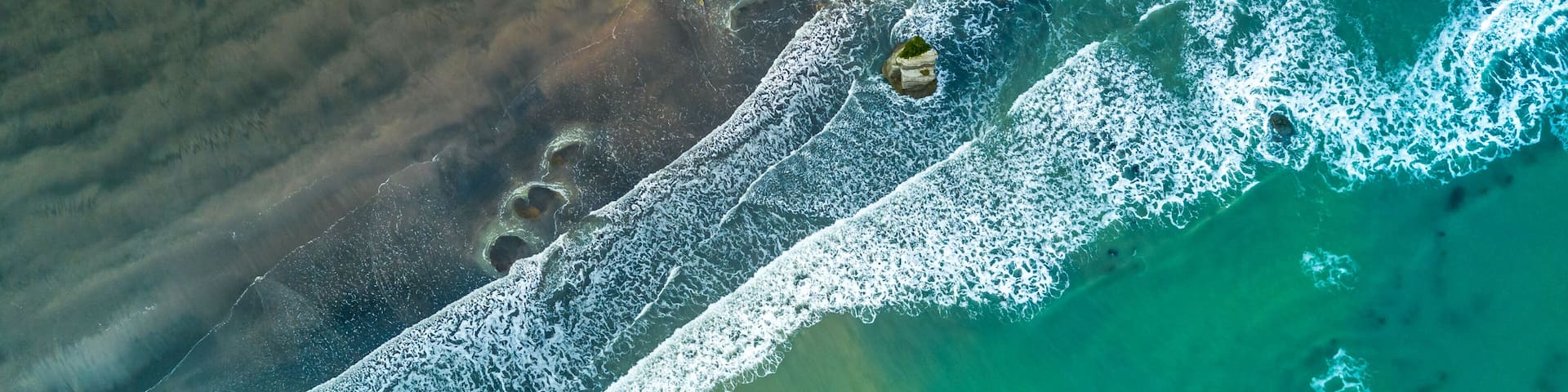 An aerial view of a beach with clear turquoise water, white waves, and rocky outcrops.
