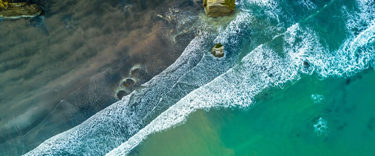 An aerial view of a beach with clear turquoise water, white waves, and rocky outcrops.