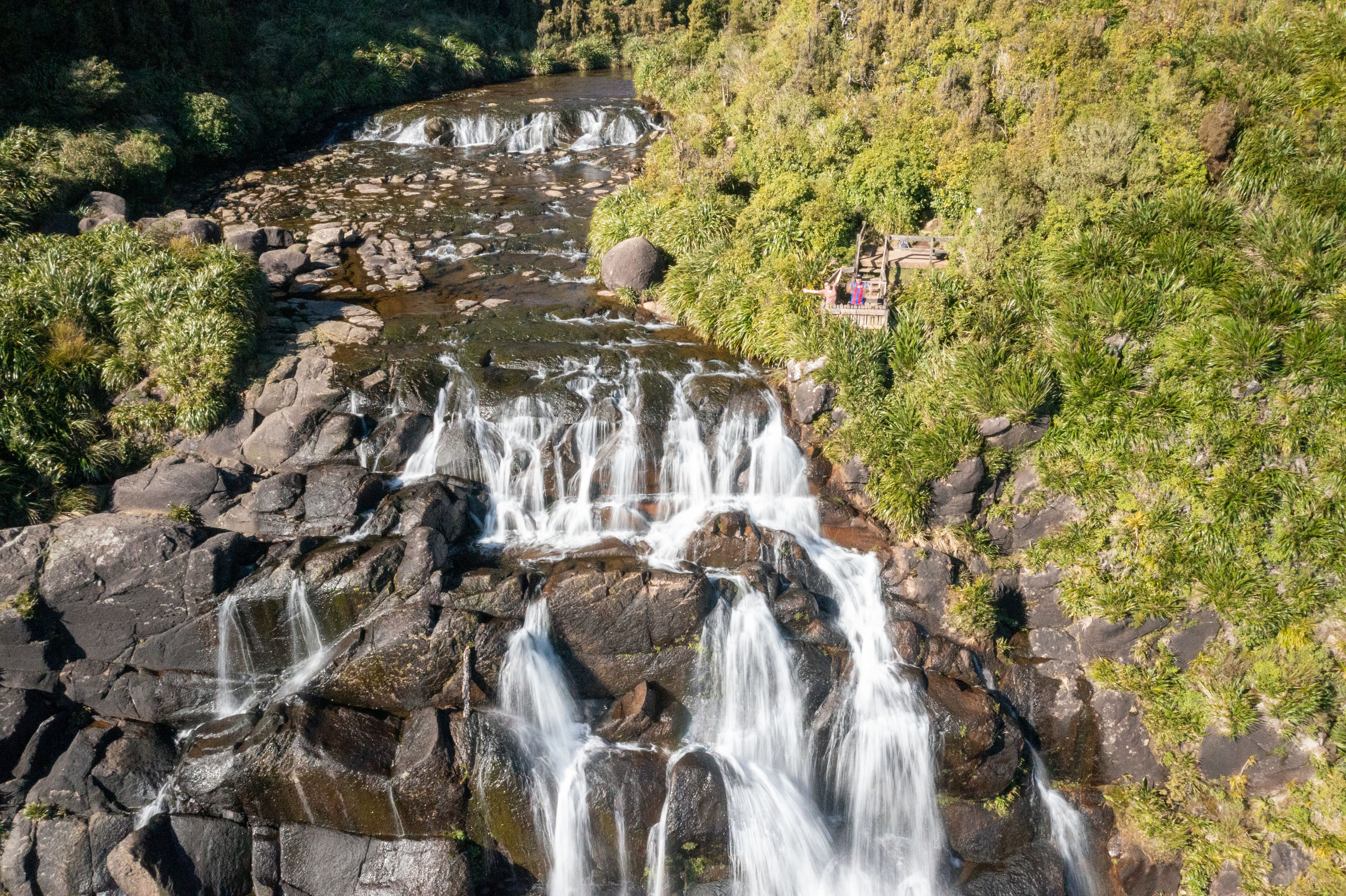 Aerial view of Wairere Falls cascading over dark rocks surrounded by lush green forest and a viewing platform in Okauia, Waikato Region, New Zealand.