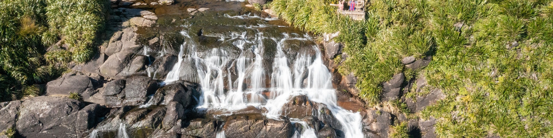 Ein mehrstufiger Wasserfall stürzt über felsiges Gelände, umgeben von üppigem Grün und einer kleinen Holzhütte.