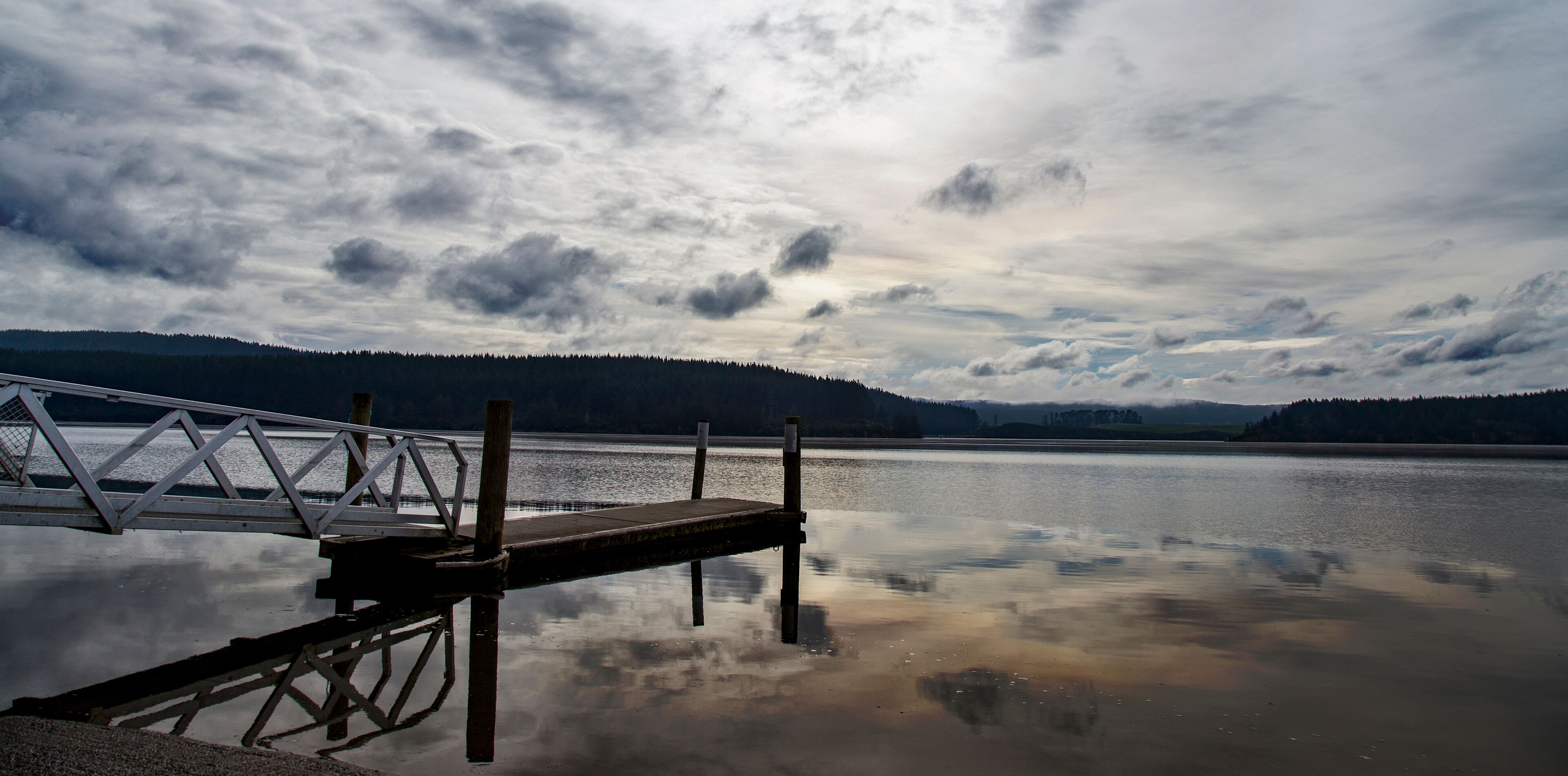 Lake Maraetai in Mangakino, Waikato district, New Zealand. Still waters, small boat jetty, reflections of cloud and sky. Moody, peaceful, reflective. Hydroelectric station, recreational activities.