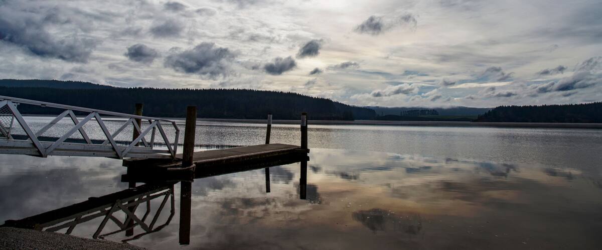 A calm lake with a wooden dock extending into the water, surrounded by forested hills under a cloudy sky.