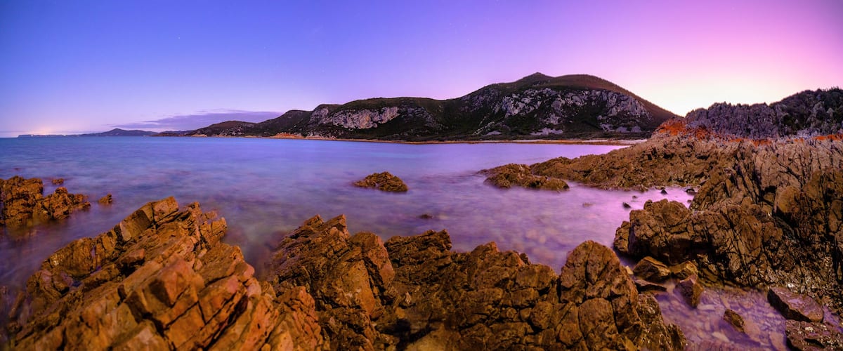 A coastal landscape with rocky shores, calm waters, and distant mountains under a twilight sky.