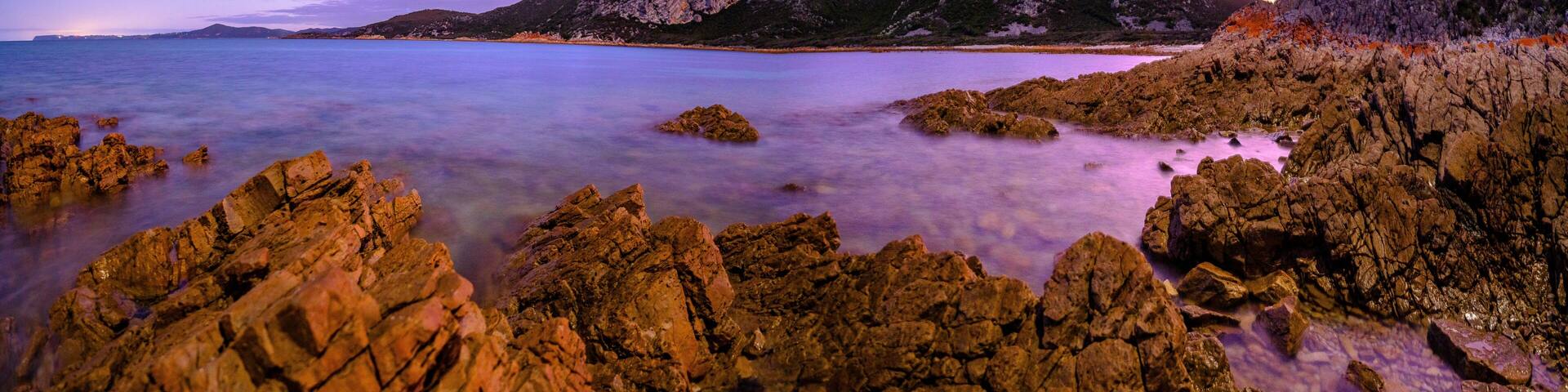 A coastal landscape with rocky shores, calm waters, and distant mountains under a twilight sky.