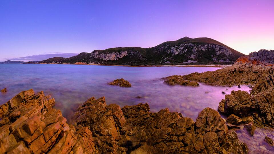A coastal landscape with rocky shores, calm waters, and distant mountains under a twilight sky.