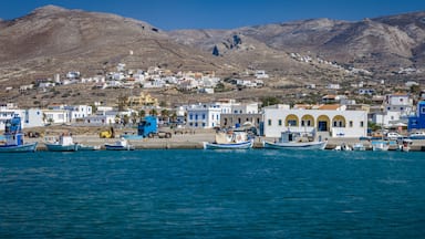 Uma vila costeira com barcos atracados ao longo da orla, edifícios com fachadas azuis e brancas, e montanhas ao fundo.