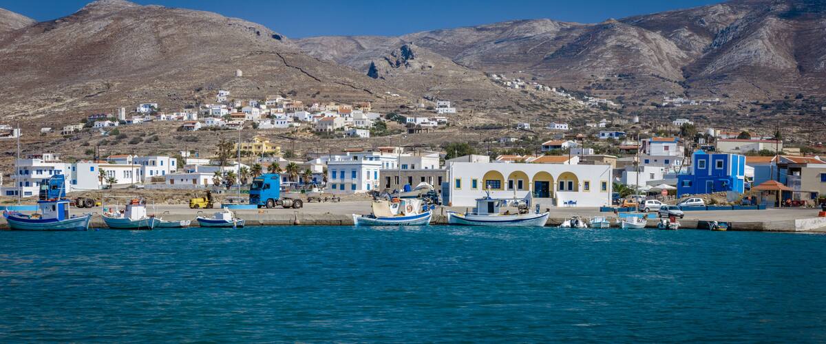 A coastal town with boats docked along the waterfront, buildings with blue and white facades, and a mountainous backdrop.