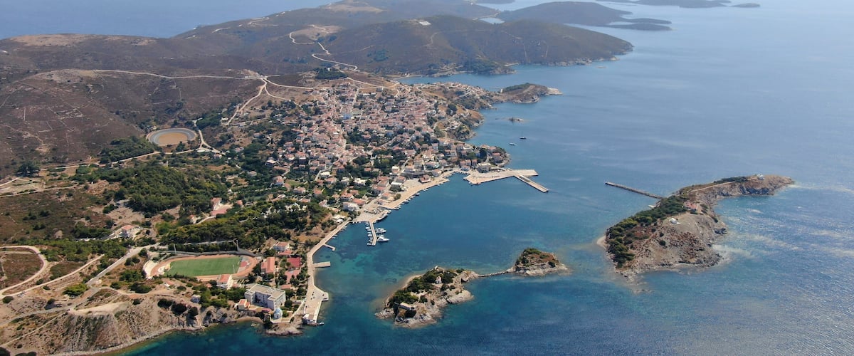An aerial view of a coastal town with a marina, surrounded by hills and the sea.
