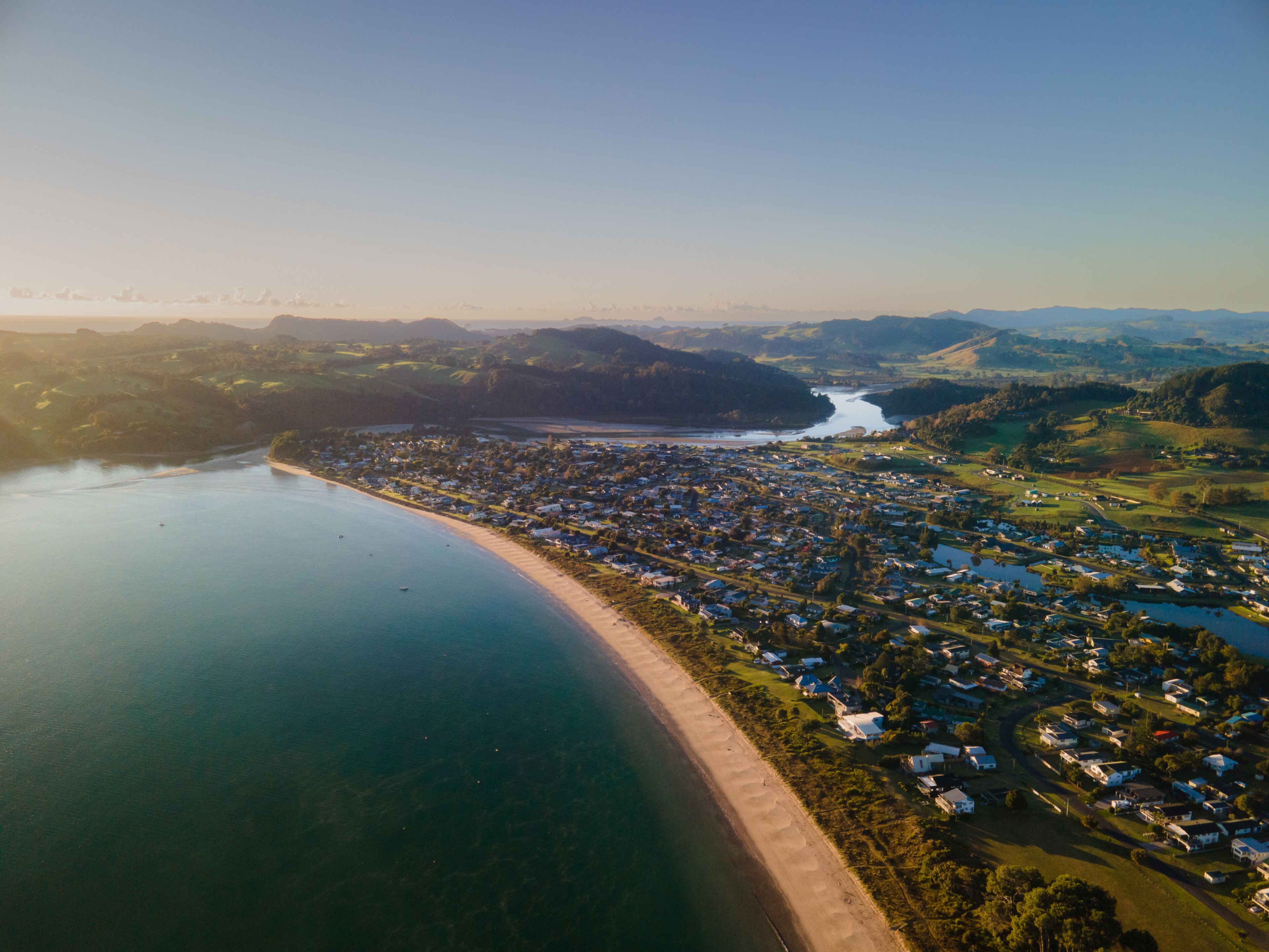 Uma vila costeira com uma praia, um rio e colinas suaves ao fundo.