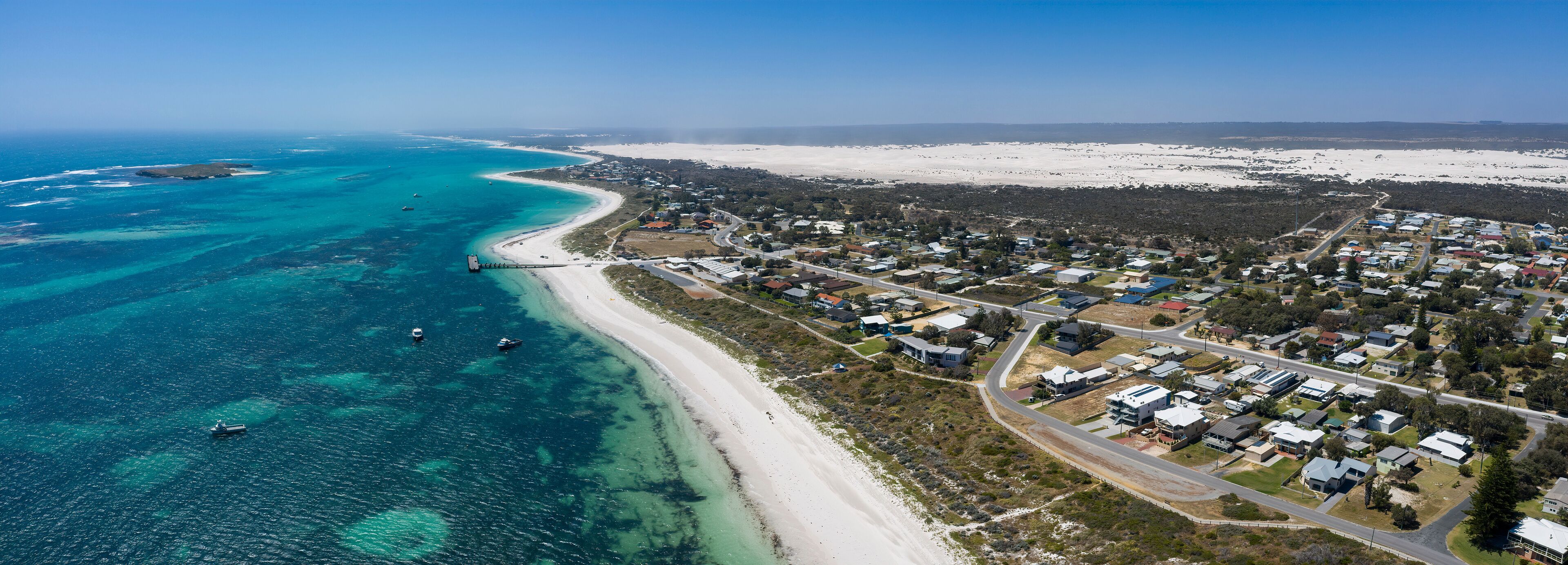 An aerial view of a coastal town with a beach, clear blue waters, and a mix of residential and natural landscapes.