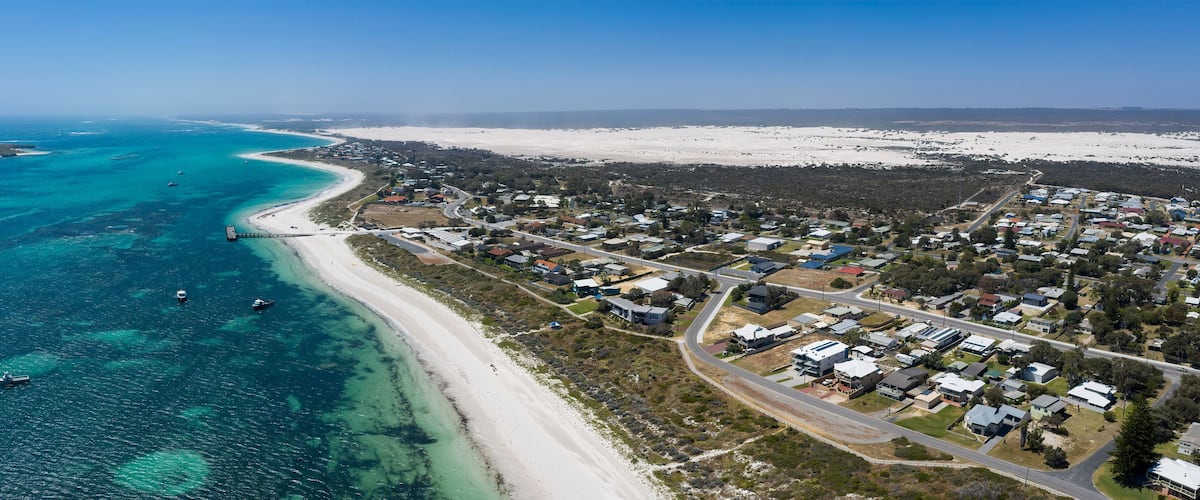 An aerial view of a coastal town with a beach, clear blue waters, and a mix of residential and natural landscapes.