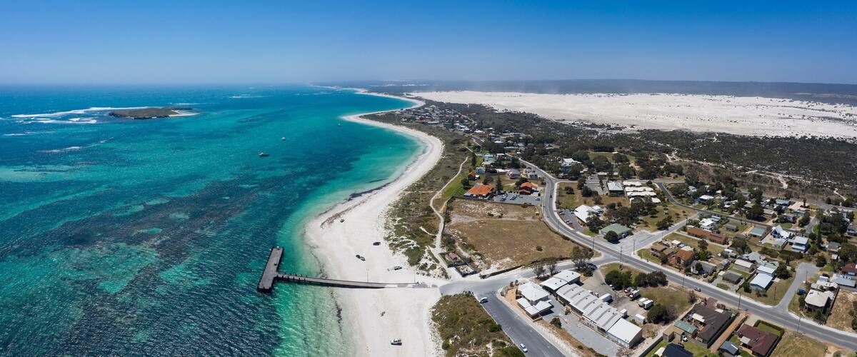 An aerial view of a coastal town with a beach, clear blue waters, and a pier.