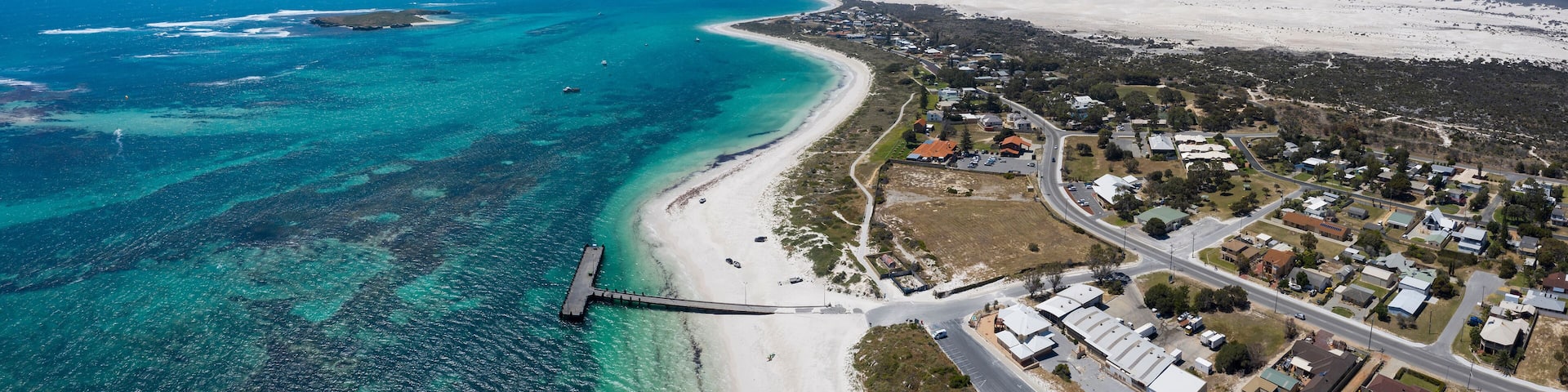 An aerial view of a coastal town with a beach, clear blue waters, and a pier.
