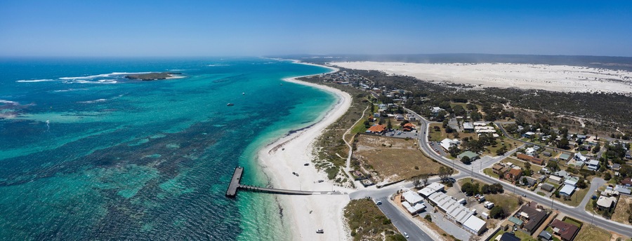 Vista aérea de un pueblo costero con playa, aguas azul turquesa y un muelle.