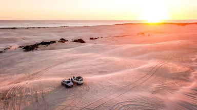 Paisagem desértica com um pôr do sol, apresentando um veículo branco e marcas na areia.