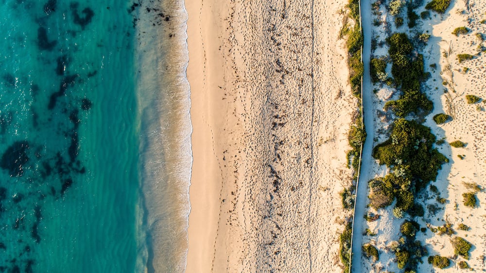 A coastal landscape with a sandy beach, clear turquoise waters, and sparse vegetation.