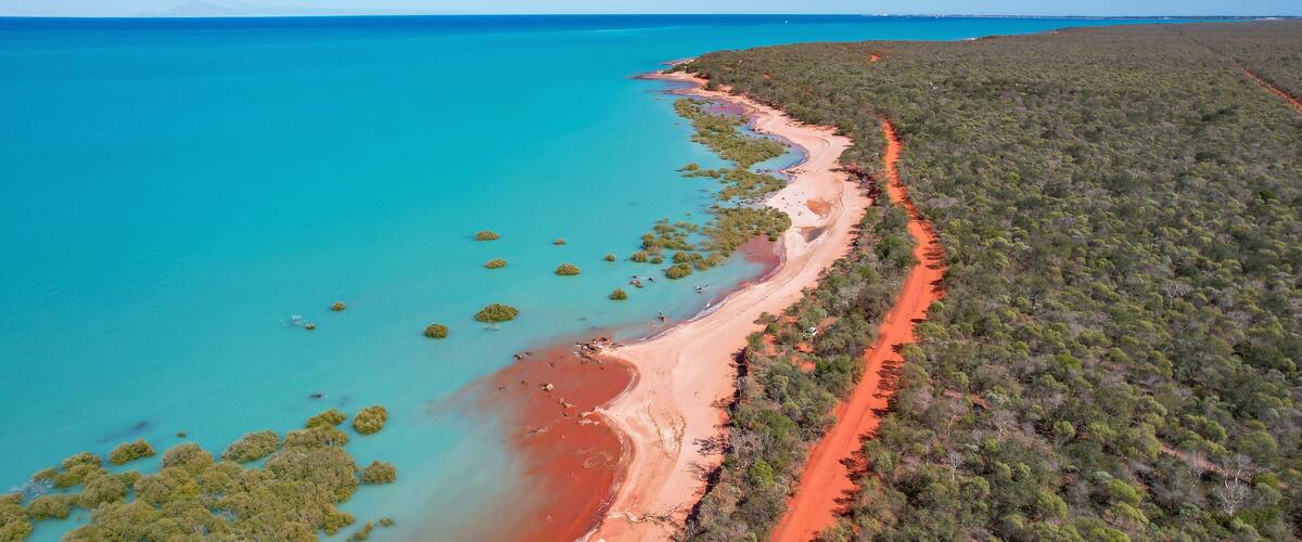 A coastal landscape with a sandy beach, clear turquoise waters, and a dirt path along the shore.