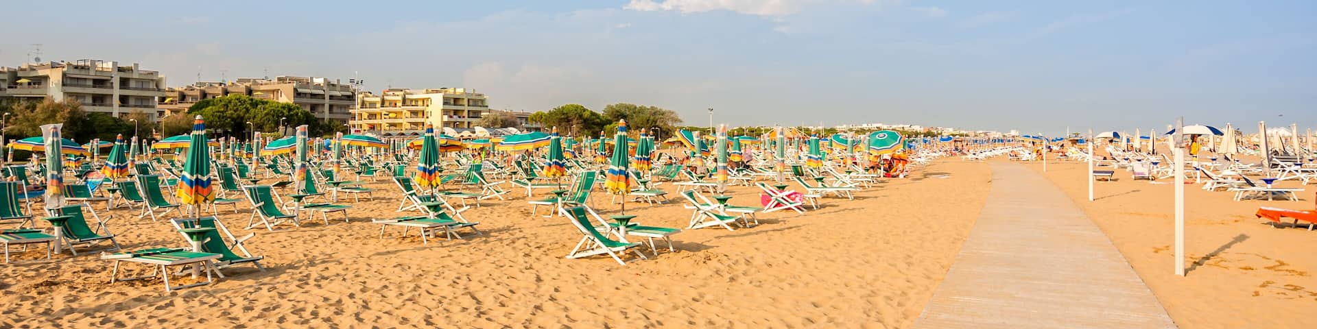 A beach with rows of green and white sun loungers and umbrellas, a sandy walkway, and buildings in the background.