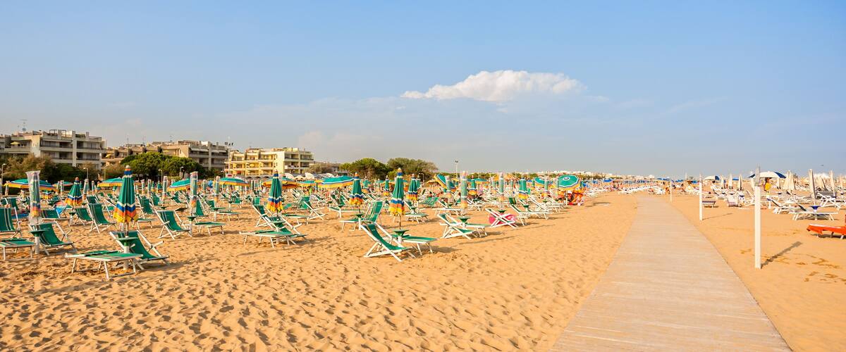 A beach with rows of green and white sun loungers and umbrellas, a sandy walkway, and buildings in the background.