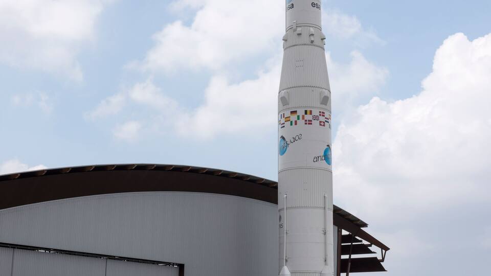 A tall rocket with a cylindrical body and a pointed top, standing on a concrete pad with a hangar in the background.