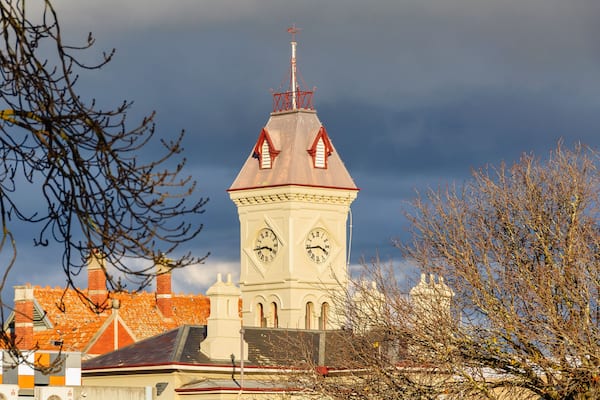 A clock tower with two faces, surrounded by buildings and bare trees against a cloudy sky.