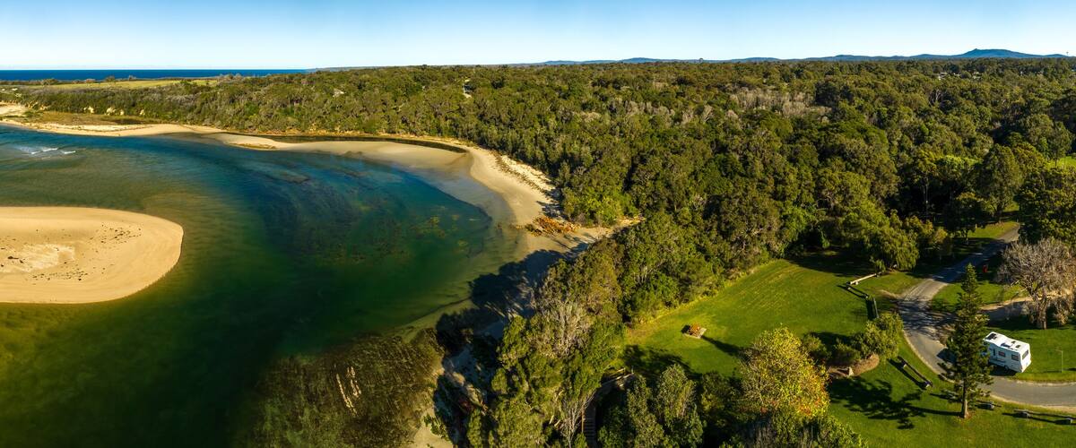 A coastal landscape with a sandy beach, clear waters, dense forest, and a road with a camper.