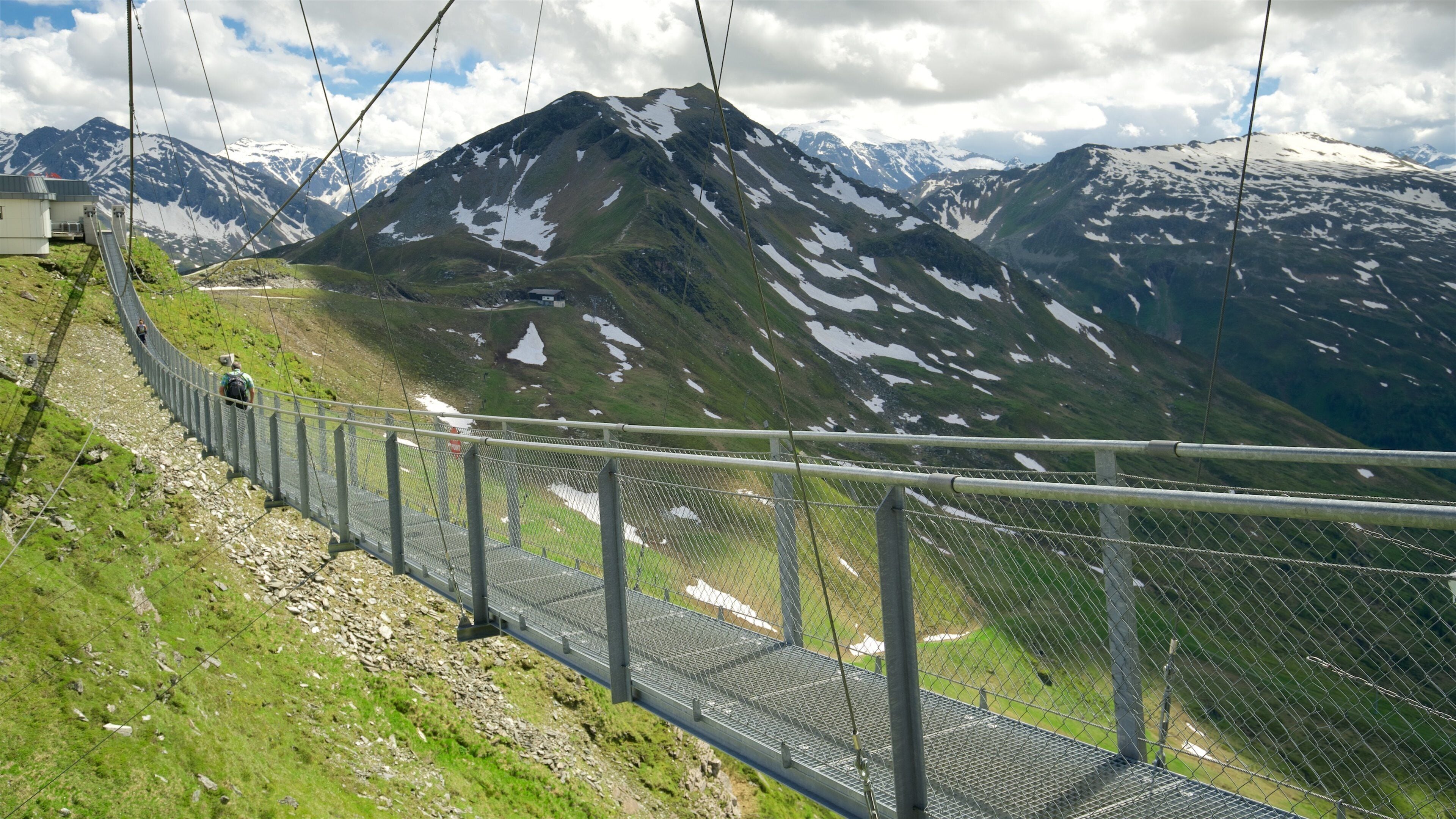 A suspension bridge with a metal fence, surrounded by mountains with patches of snow.