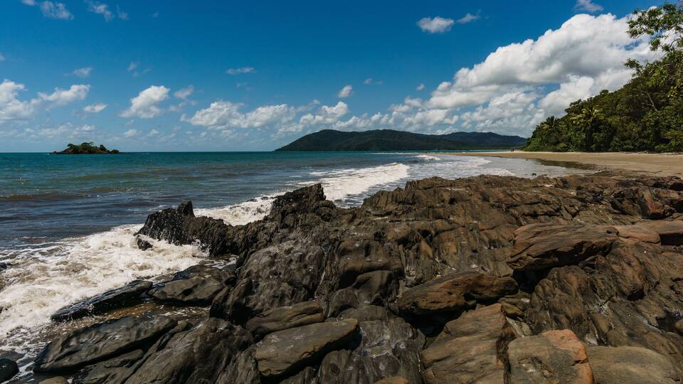 A rocky shoreline with waves crashing onto the rocks, a sandy beach, and a distant island under a clear blue sky.