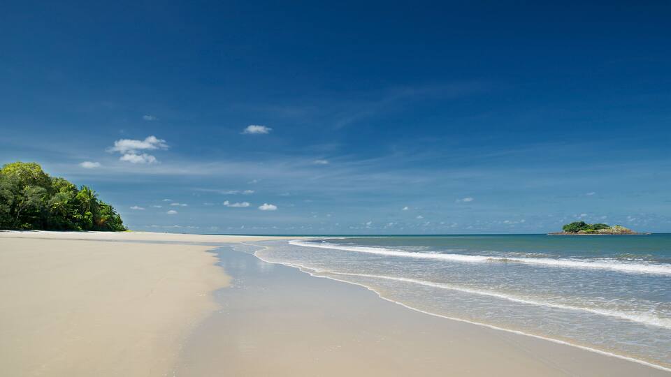 A beach with clear blue water, a sandy shore, and a small island with trees in the distance.