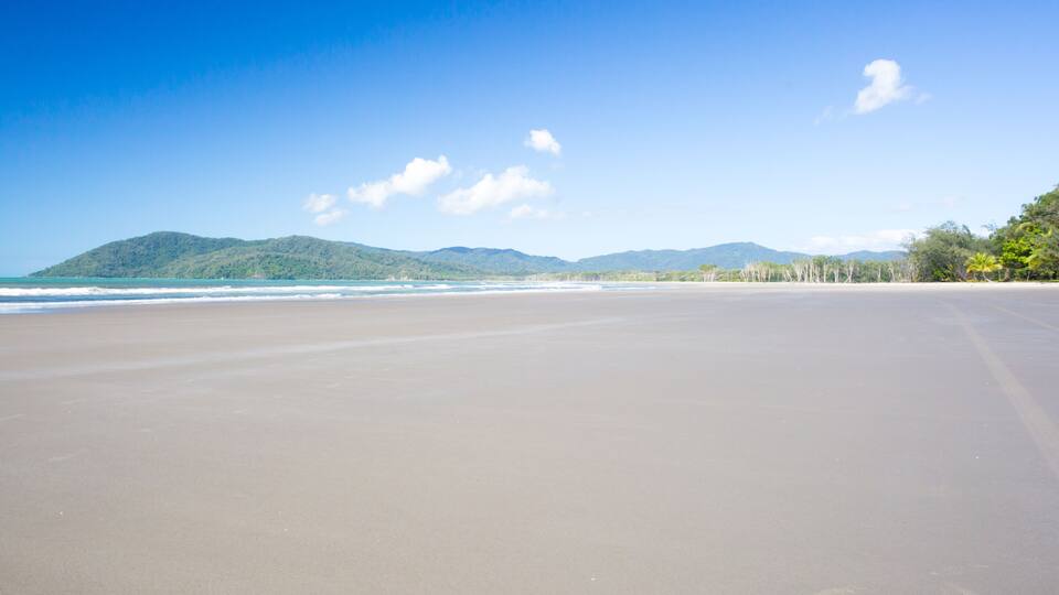 A wide sandy beach with gentle waves, a clear blue sky, and distant green hills.