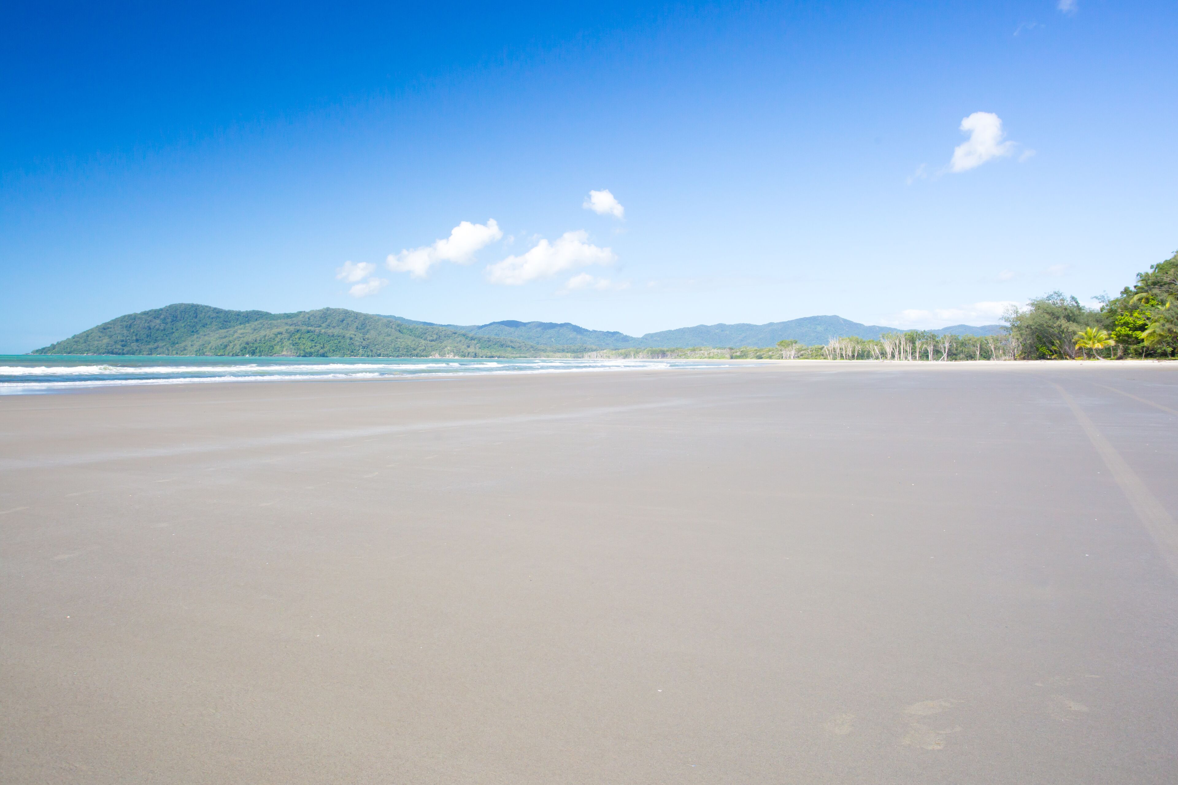A wide sandy beach with gentle waves, a clear blue sky, and distant green hills.