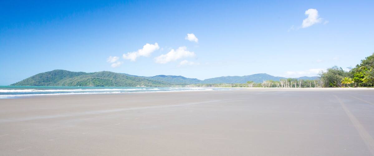 A wide sandy beach with gentle waves, a clear blue sky, and distant green hills.