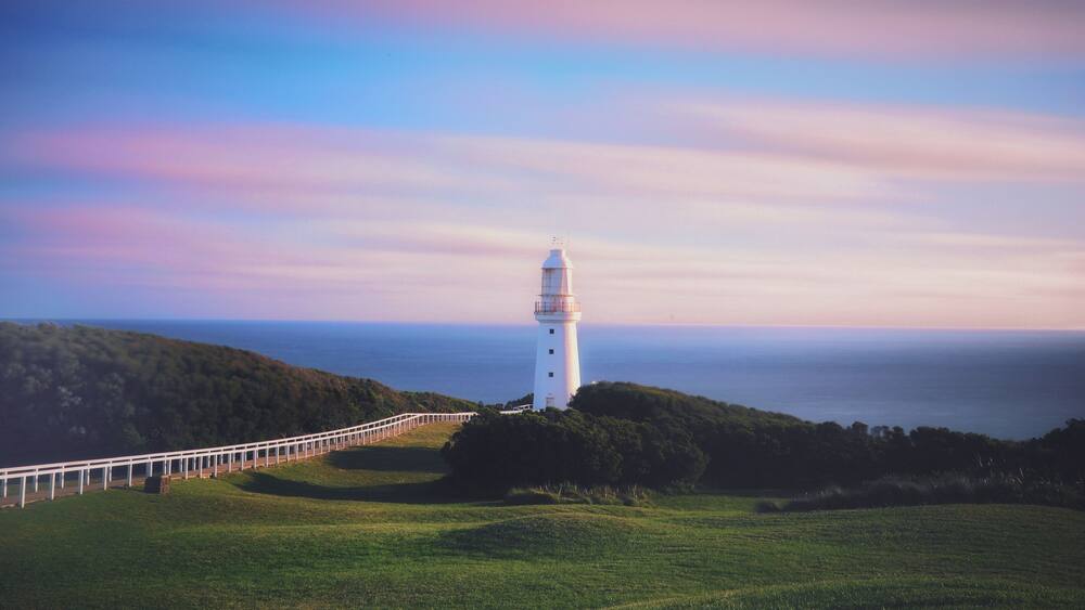 A white lighthouse on a grassy hill with a white fence leading up to it.