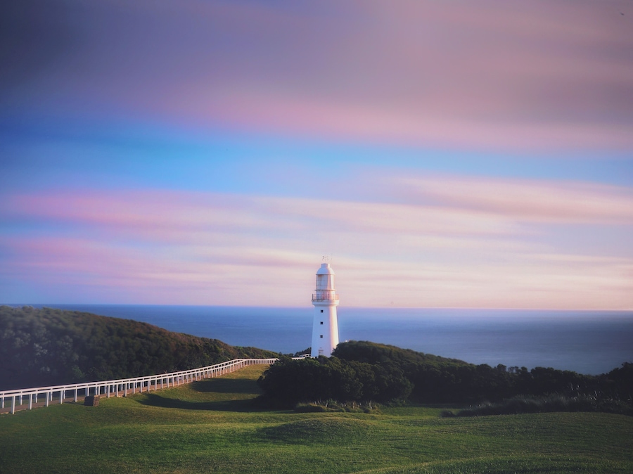 Un faro blanco sobre una colina cubierta de hierba, con una cerca blanca que conduce hasta él.
