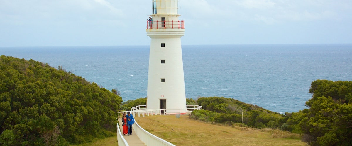 A white lighthouse with a red railing, surrounded by greenery and overlooking the ocean.