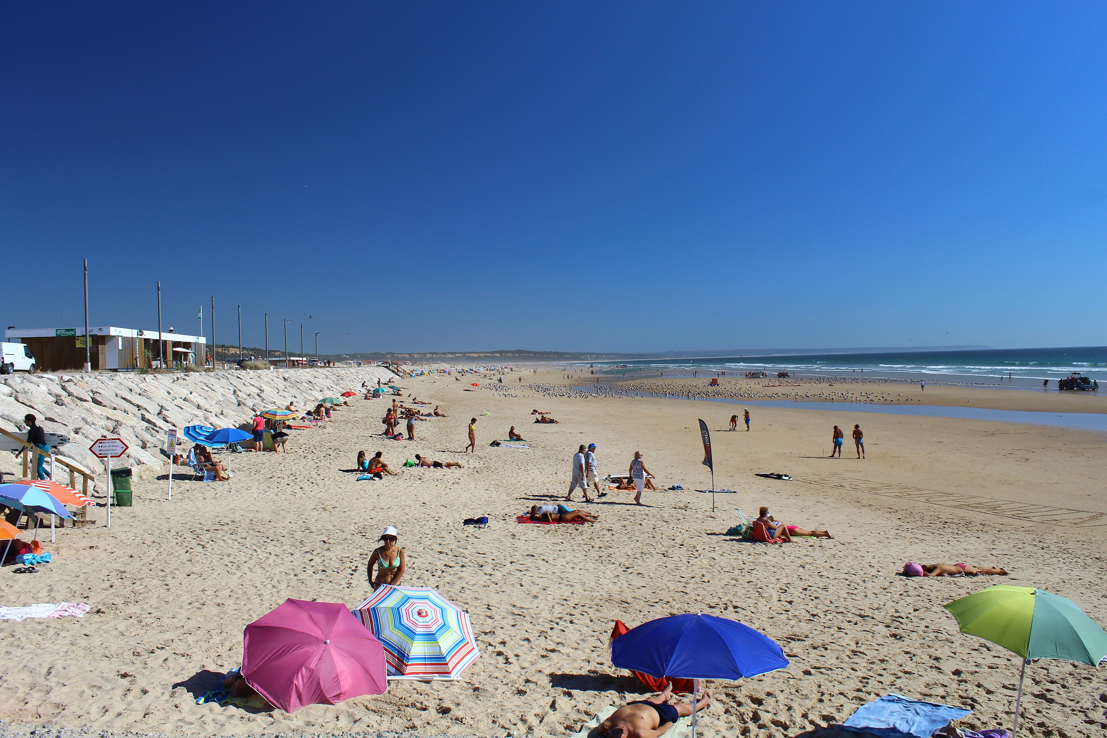 A sandy beach with people sunbathing and walking, colorful umbrellas, and a clear blue sky.