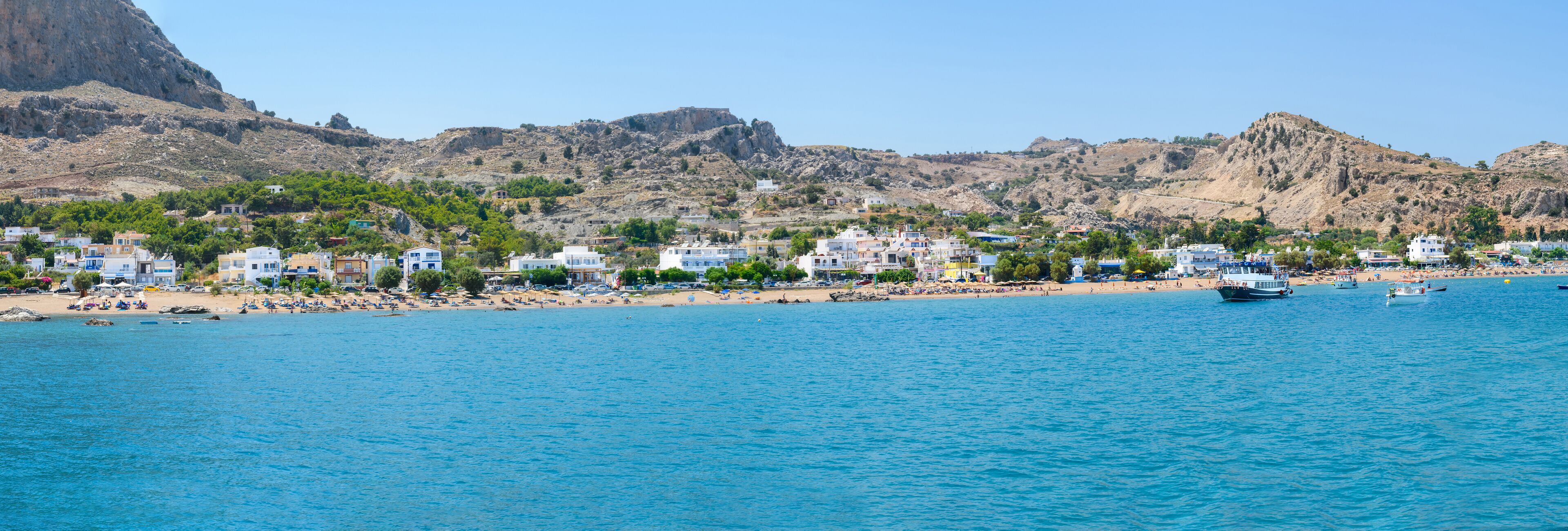 A coastal town with buildings along the beach, a rocky mountain in the background, and a clear blue sea.