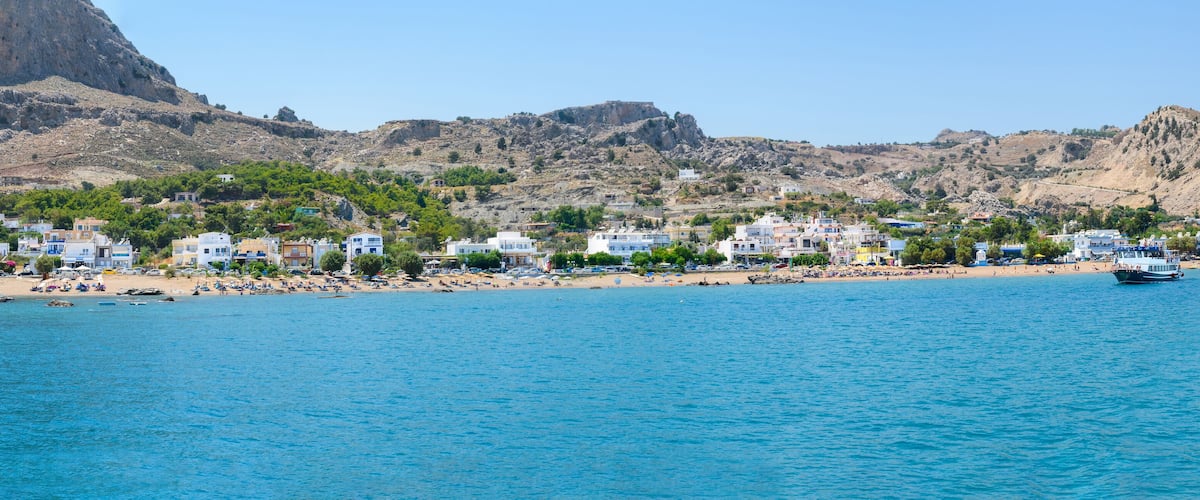 A coastal town with buildings along the beach, a rocky mountain in the background, and a clear blue sea.