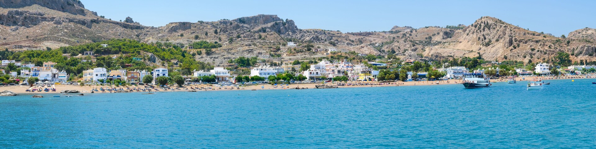 A coastal town with buildings along the beach, a rocky mountain in the background, and a clear blue sea.