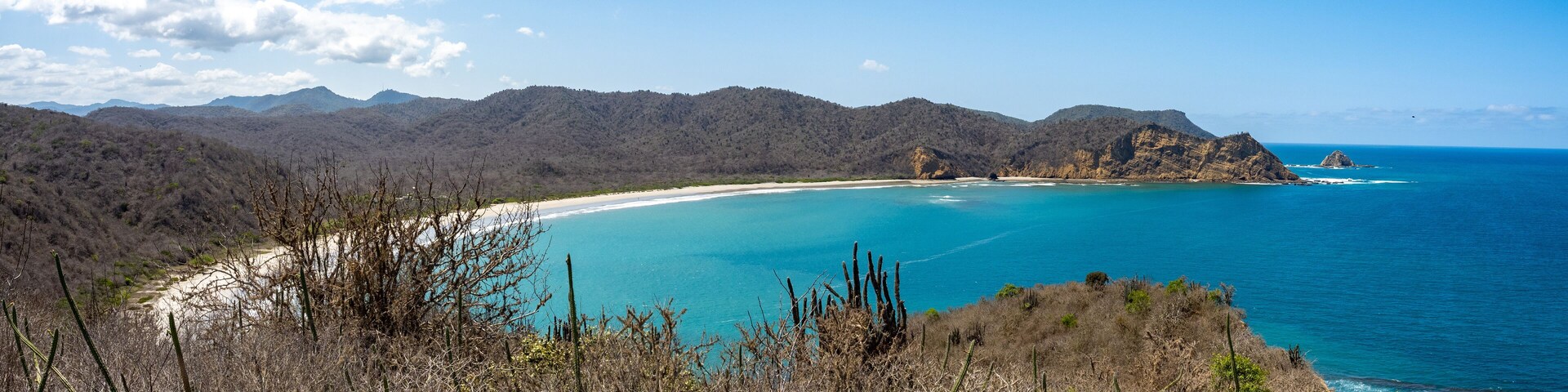 Un paisaje costero con una playa, aguas azules cristalinas y colinas boscosas.