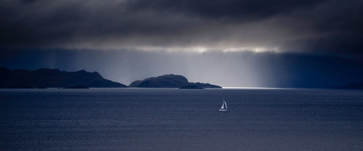 A sailboat on calm waters with a mountainous island in the distance under a cloudy sky.