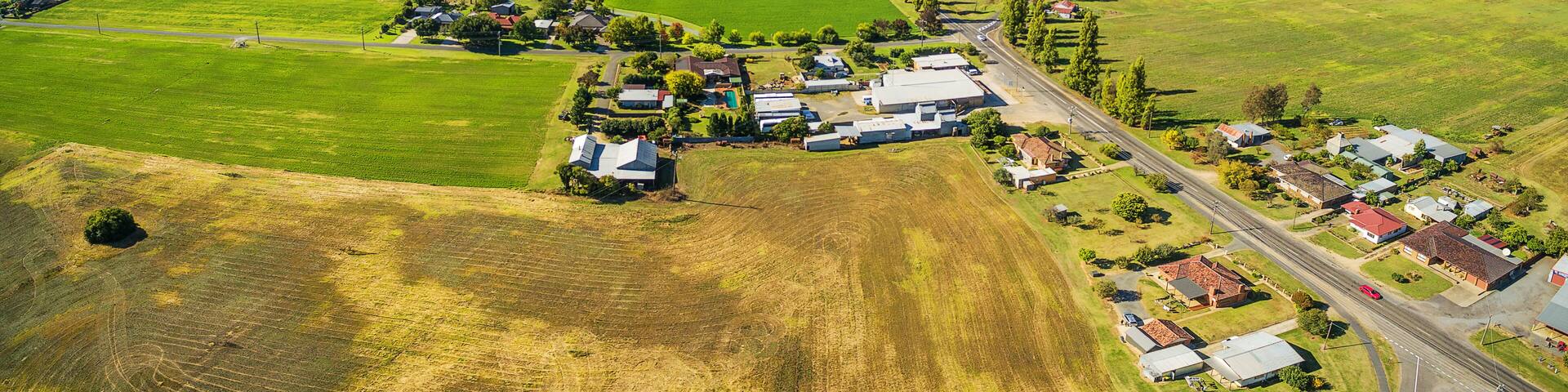 An aerial view of a rural town with green fields, houses, and mountains in the background.